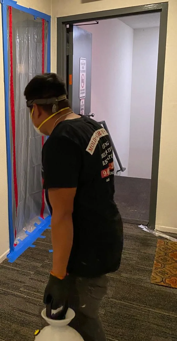 Technician in a respirator carries a pump sprayer past a doorway sealed with poly sheeting, tape, and a zipper entry.