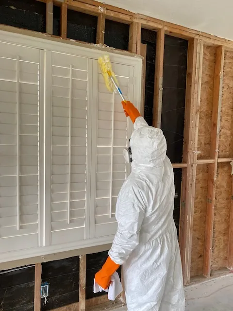 Technician in a hooded Tyvek coverall and respirator cleaning above plantation shutters in a room stripped to the studs.